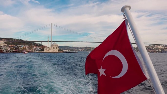 船的POV土耳其国旗在海上与Ortakoy清真寺和博斯普鲁斯桥在多云的天空背景