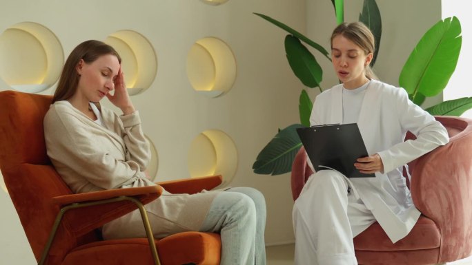 一位忧心忡忡的年轻病人坐在心理医生的预约诊室里谈论心理问题，一位细心的女医生倾听并做笔记