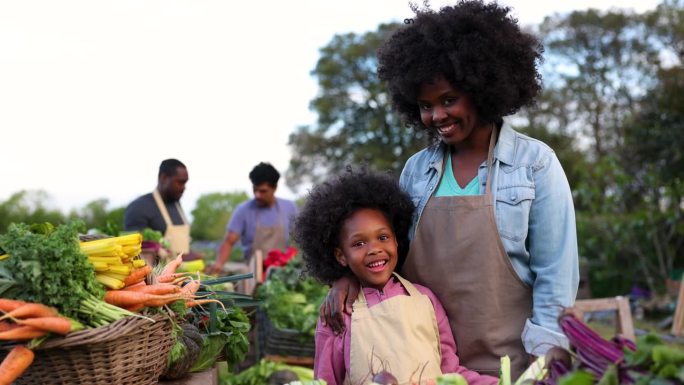 可爱的黑人小女孩欢迎顾客从当地农场购买新鲜蔬菜，两人都对着镜头微笑——背景是杂工