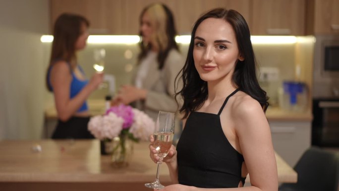 在前景中，一个美丽的白人女孩喝着玻璃杯里的白起泡酒，背景是她的朋友们在一个现代公寓的厨房里聊天。
