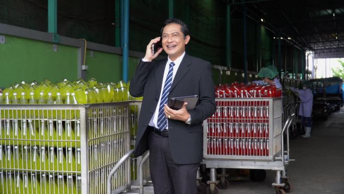 亚洲资深商务人士站在饮料仓库工厂边打电话，联系商家订货