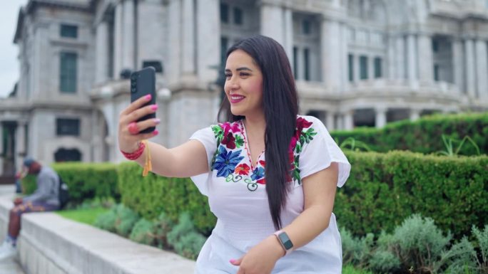 快乐的年轻西班牙女子坐在墨西哥城贝拉斯美术宫前，在日光下享受着地理位置，穿着传统服装，用智能手机自拍