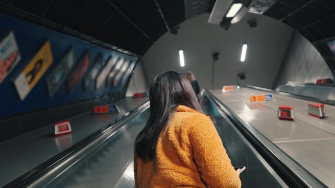在英国伦敦地铁站的自动扶梯上，亚洲女游客一边用手机一边环顾四周的慢镜头
