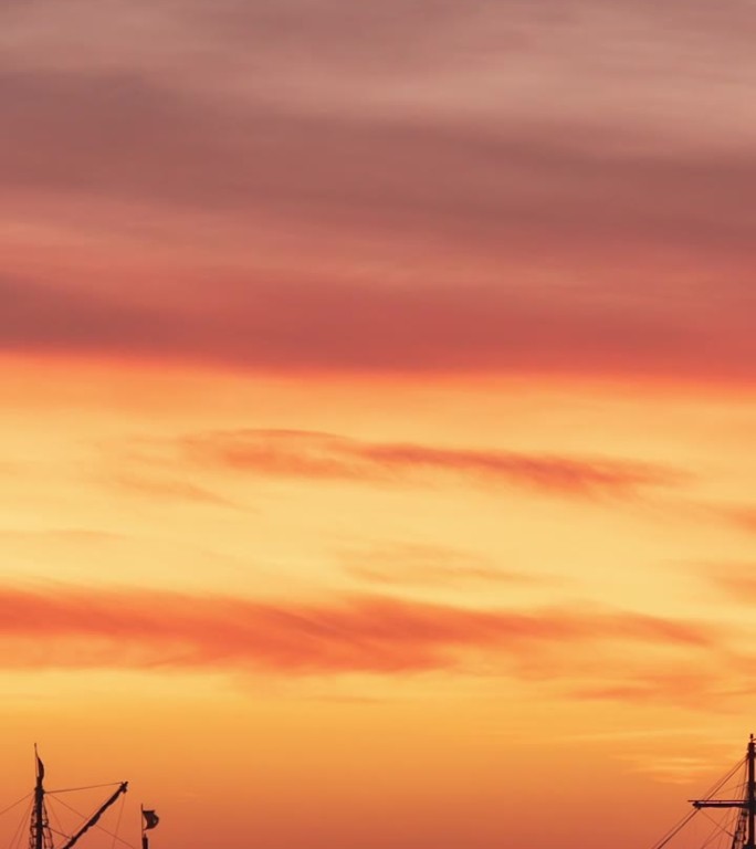 垂直视频。古老帆船的高桅杆和顶上展开的破烂旗帜，背景是冉冉升起的太阳和火红的云。延时。