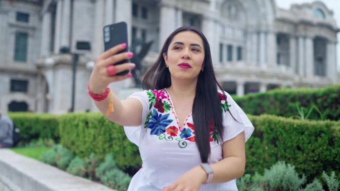 快乐的年轻西班牙女子坐在墨西哥城贝拉斯美术宫前，在日光下享受着地理位置，穿着传统服装，用智能手机自拍