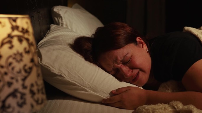 愁眉苦脸的女人躺在床上的枕头上，在黑暗的卧室里痛苦地哭泣。夜临睡前歇斯底里，泣不成声。
