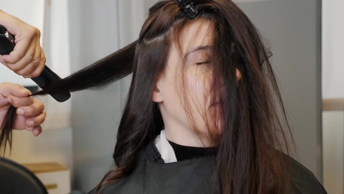 发型师用电卷发棒给女孩做头发。女人卷发，造型。为假期做准备。在豪华美容院，女理发师正在用卷发棒处理一