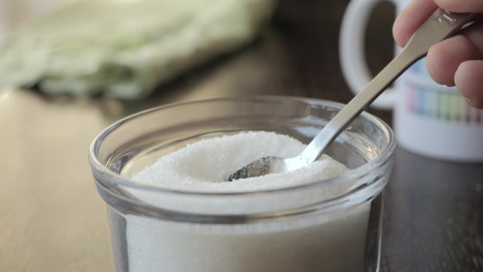 用勺子舀清白糖的慢动作。从玻璃糖碗中取出一茶匙糖。甜食上瘾的垃圾食品。不健康饮食的概念，糖尿病，肥胖