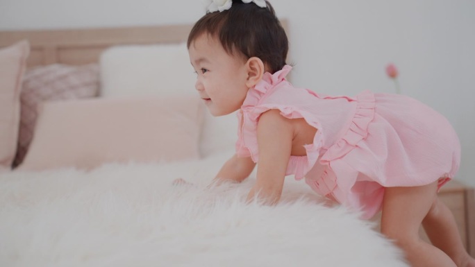 可爱的亚洲女婴穿着可爱的粉色连衣裙，在卧室里爬上床，试图够到床上的东西。