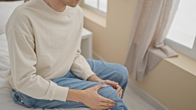 一头卷发的年轻人坐在家里的床上，抱着膝盖，表现出疼痛或不适，背景是中性的装饰。