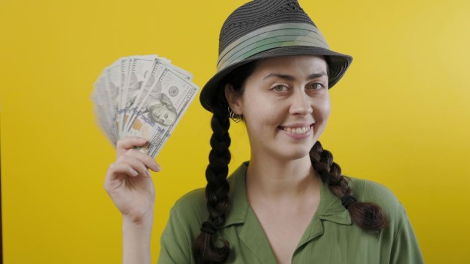 财富和投资。一个戴着草帽的快乐的白人妇女的肖像，展示了一个美元的扇子。黄色背景。彩票中奖的概念