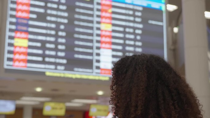 一头短卷发的黑人妇女正在机场的屏幕上查看航班号、登机时间和登机门号码，以便登机旅行或学习。