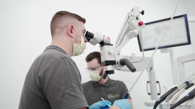 现代牙科诊所，男牙医与助手用显微镜操作病人牙齿。医生清理牙根管，烧灼牙神经。牙髓医生治疗病人的根管。