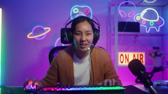 快乐的亚洲女性玩家戴着耳机比赛玩视频游戏与电脑在客厅彩色霓虹灯在线
