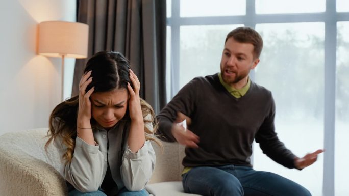 一对白人夫妇坐在客厅的沙发上，因为意见不合而争吵，男人对女人大喊大叫，女人遭受丈夫的心理虐待