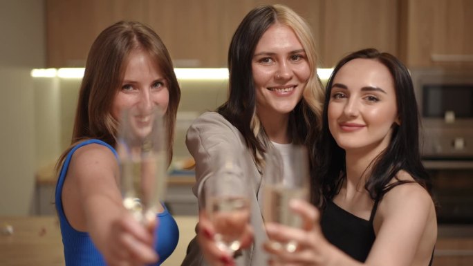 三个女孩站在一间现代公寓的厨房里，手里拿着几杯白起泡酒。女孩们对着镜头拿出笛子，喝着酒，微笑着看着镜