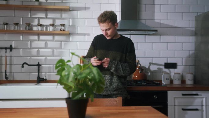 年轻帅气的小伙子把茶壶放在现代厨房的炉子上，从口袋里掏出手机，坐在厨房的长凳上查看短信