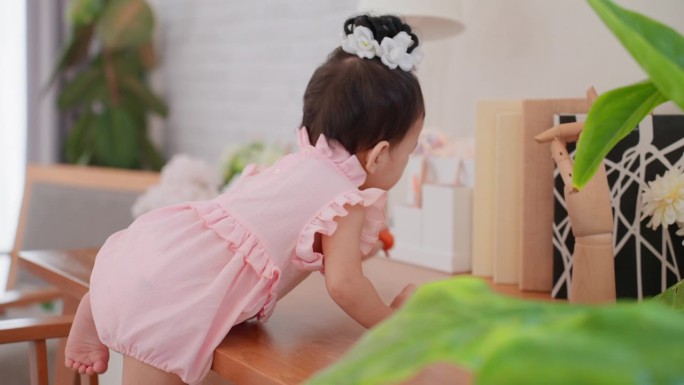可爱的亚洲女婴穿着可爱的粉色连衣裙，爬上她家客厅的高桌上，试图够到东西。