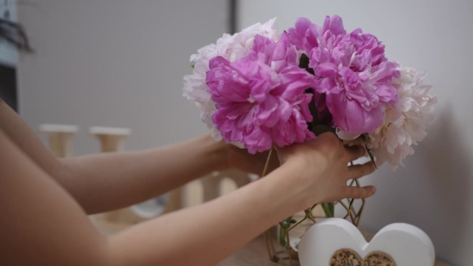 在一间公寓的现代客厅里，一个女孩把粉红色和白色的牡丹放在木制床头柜上的玻璃花瓶里。一个留着黑色长发的