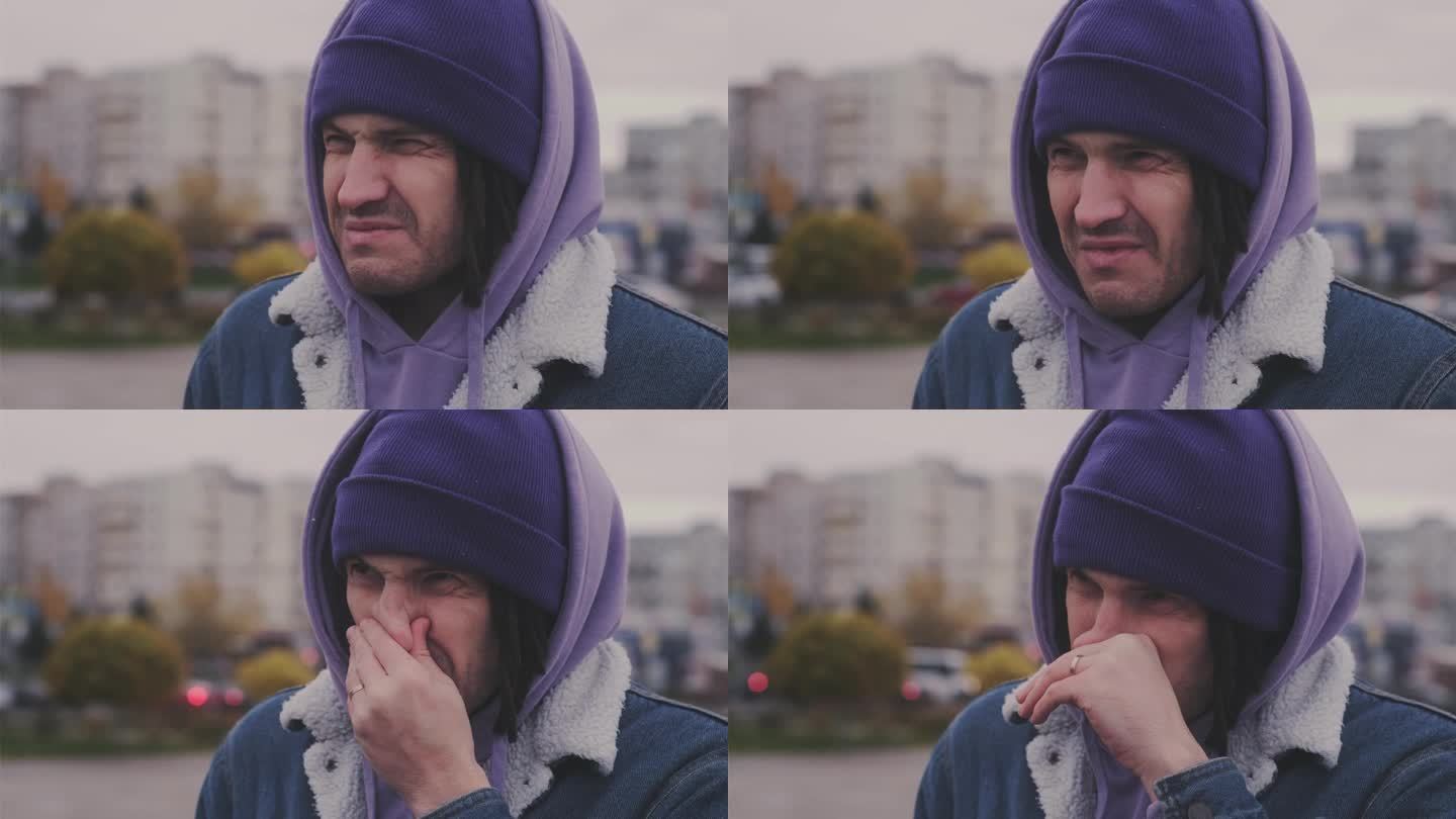 一名男子站在城市街道上，在寒冷的天气里抽泣。冻僵的人穿着暖和的衣服，流着鼻涕，擦着鼻子。
