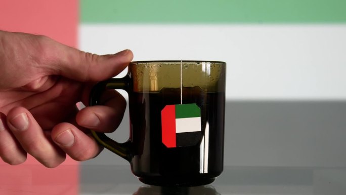 一个男人拿着一杯茶的特写镜头，茶包上有阿联酋国旗的标签，从玻璃桌子上拿下来，对面是同样模糊的国旗