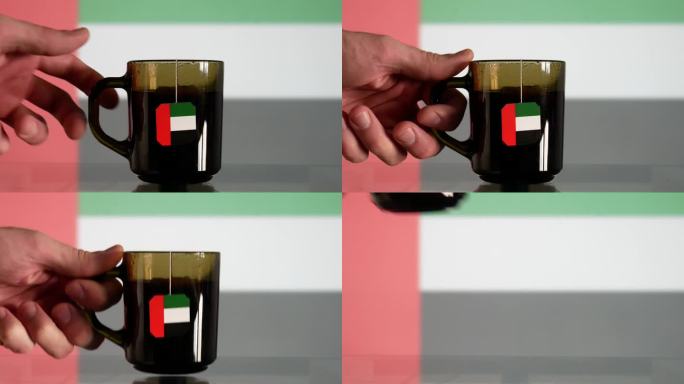 一个男人拿着一杯茶的特写镜头，茶包上有阿联酋国旗的标签，从玻璃桌子上拿下来，对面是同样模糊的国旗