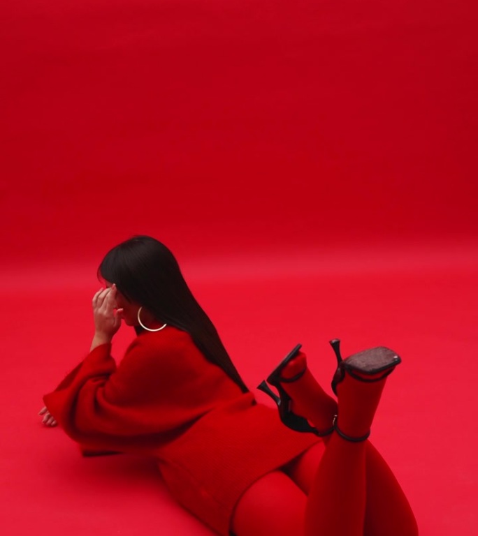 亚洲女模特身穿红色毛衣和连裤袜，趴在工作室红色背景的地板上。艺妓穿着红色的衣服。垂直视频