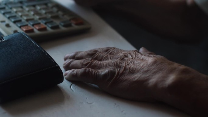 一个领养老金的人正在用计算器计算开支，他旁边的桌子上放着一个钱包。老年财务会计。养老金储蓄，红利，养