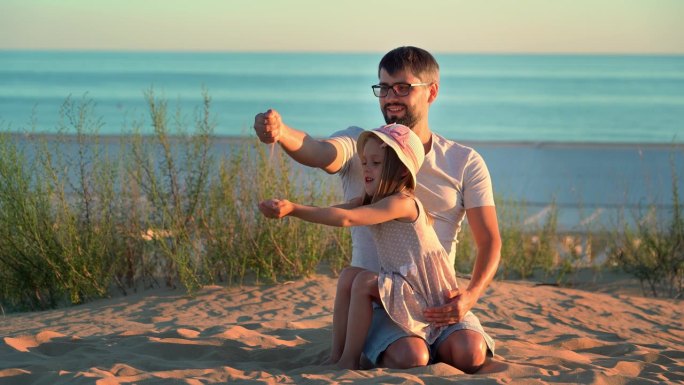 夕阳下，父亲和女儿在沙滩上玩沙子，背景是蓝色的大海