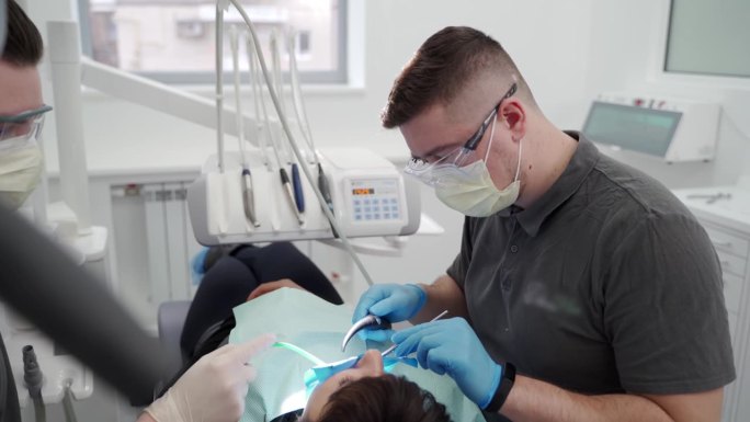 在现代牙科诊所工作的牙医和助理。医生取出牙齿上的结石和硬菌斑，助手手持唾液喷射器。口腔医生为病人做手