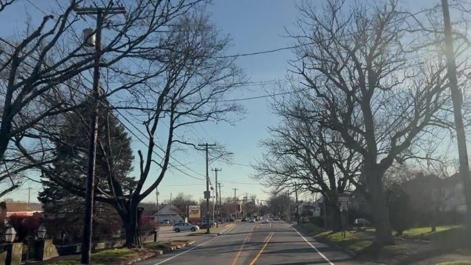 在一个晴朗的日子里，天空中有淡淡的云，汽车驶过一条两旁是树木、小房子和当地企业的街道