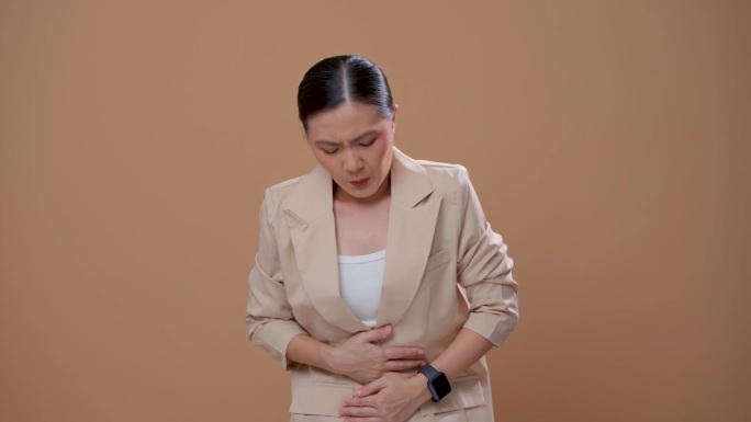一名亚洲妇女胃痛，独自站在米色背景上。