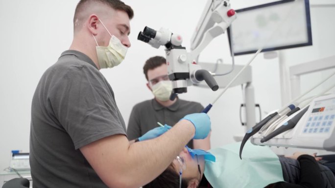 现代牙科诊所男牙医与助手用显微镜操作病人牙齿。医生清理牙根管，烧灼牙神经。牙髓医生治疗病人的根管。
