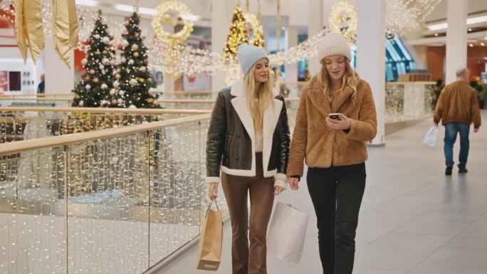圣诞期间，快乐的年轻女性朋友们穿着时髦的衣服，提着购物袋，在商场里玩着智能手机