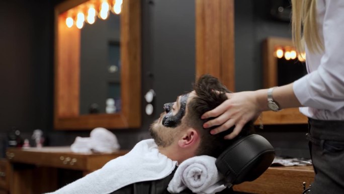 男子斜倚在理发椅上，接受女理发师的黑炭面膜护理。水疗服务，男士美容在现代沙龙例行。客户在男士美容过程