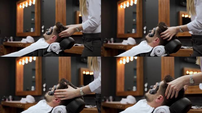 男子斜倚在理发椅上，接受女理发师的黑炭面膜护理。水疗服务，男士美容在现代沙龙例行。客户在男士美容过程