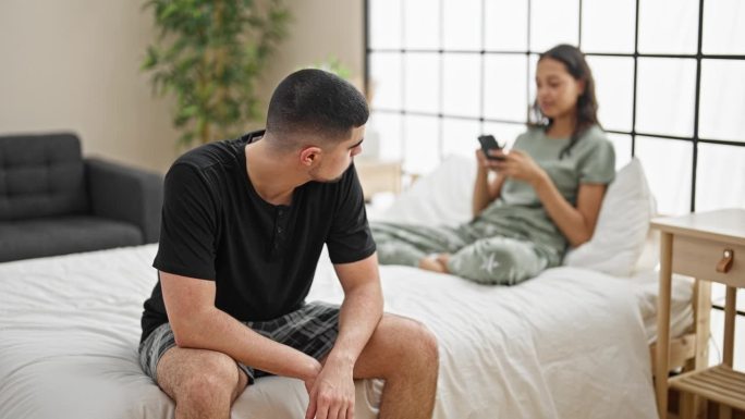 一对美丽的夫妇在卧室里意见不合，坐在床上，一边用智能手机一边表达矛盾，一个引起严肃表情的问题