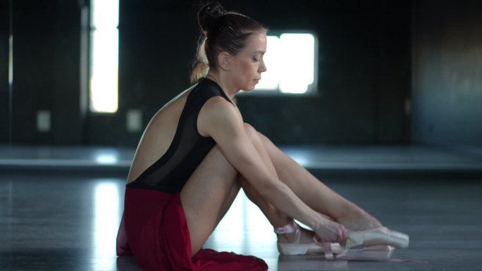 芭蕾舞女演员坐在编舞大厅的木地板上，穿上足尖鞋。概要文件。