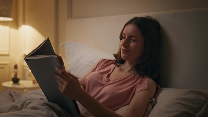平静的女性在晚上拿着书的特写。微笑的女孩读小说休息