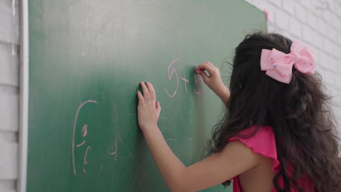可爱的多种族小学女生在黑板上写加法数学题。在一所小学的教室里，一个四岁的小女孩在黑板上展示她的数学技
