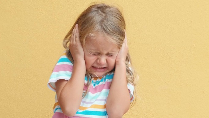 不开心悲伤愤怒的小女孩闭上眼睛捂住耳朵被嘈杂的噪音所困扰，孤立的黄色工作室背景。孩子的情绪。孩子不喜