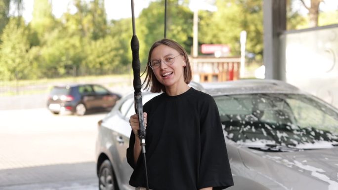 微笑的年轻迷人的女人有乐趣洗车枪，使用相机拍摄。女孩正在自助洗车店洗车。汽车清洁工具。