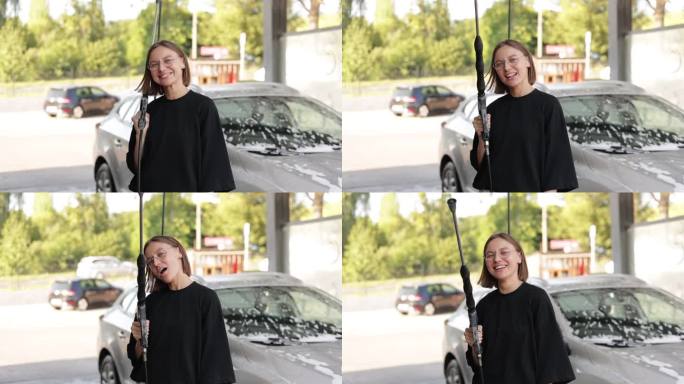 微笑的年轻迷人的女人有乐趣洗车枪，使用相机拍摄。女孩正在自助洗车店洗车。汽车清洁工具。