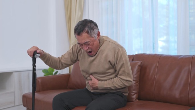 一位老人肚子疼，拄着拐杖坐在家里客厅的沙发上。