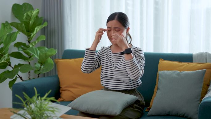 一位亚洲妇女坐在家里客厅的沙发上，眼睛酸痛。