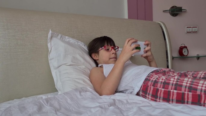 视力差的十几岁的孩子戴着眼镜眯着眼睛躺在床上看屏幕