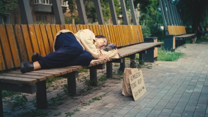 无家可归的老年女性躺在街上的长椅上，地上有帮助的标志，穷困潦倒