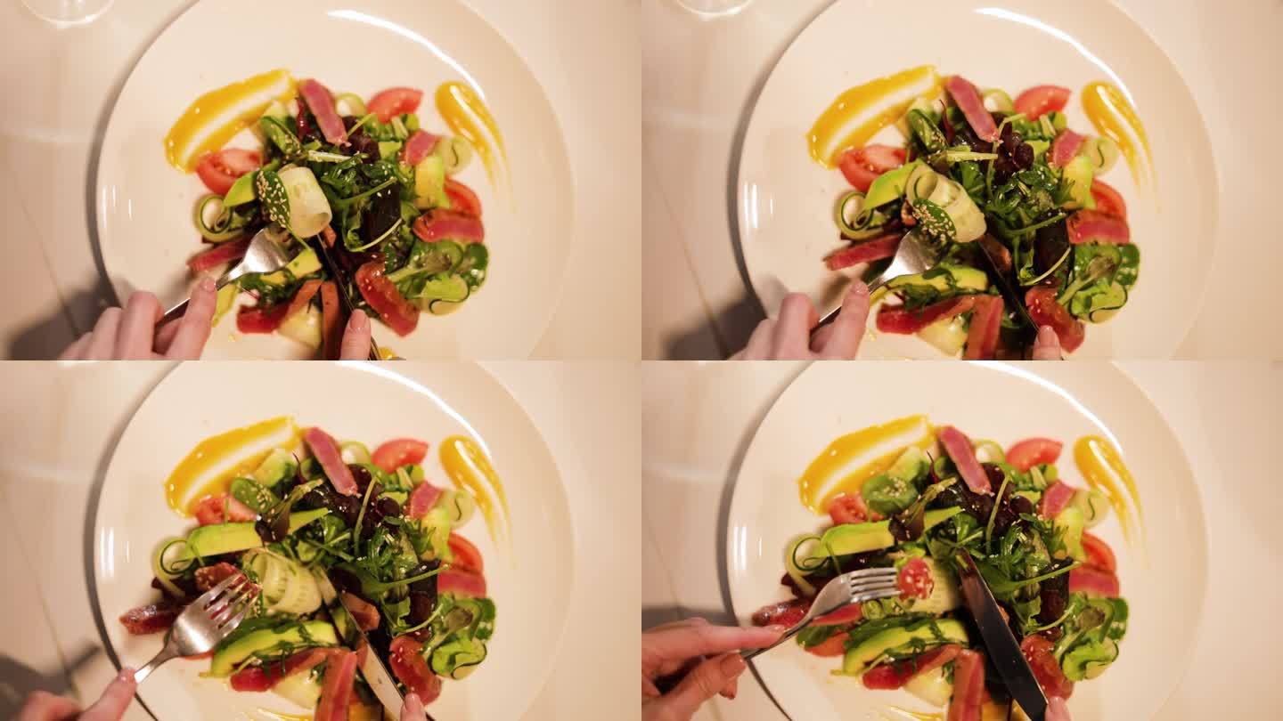 在餐厅里，顾客面前的大盘子里有蔬菜、蔬菜和火腿的有机混合沙拉。女性双手用餐具切割食材，舒适优雅。
