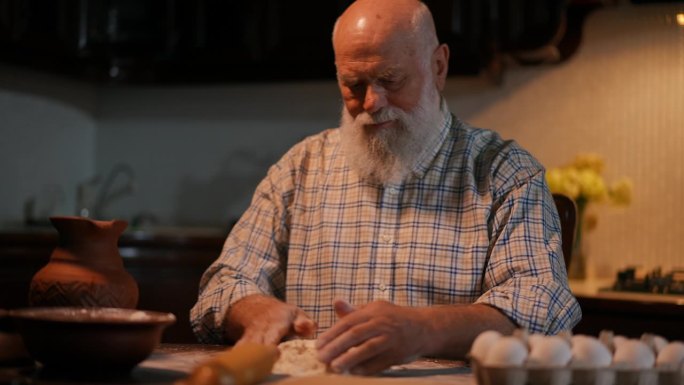 一个留着胡子的老人坐在厨房的桌子旁揉面。一名男子将陶罐中的水倒在面粉上并用手揉面。特写镜头。
