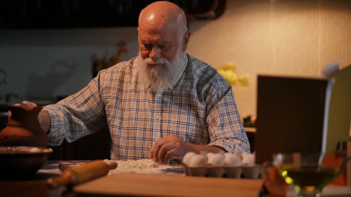 一位留着胡子的老人坐在餐桌旁，看着架子上的一本食谱。这个人从一个陶罐里往面团里加水，用手揉面，然后看